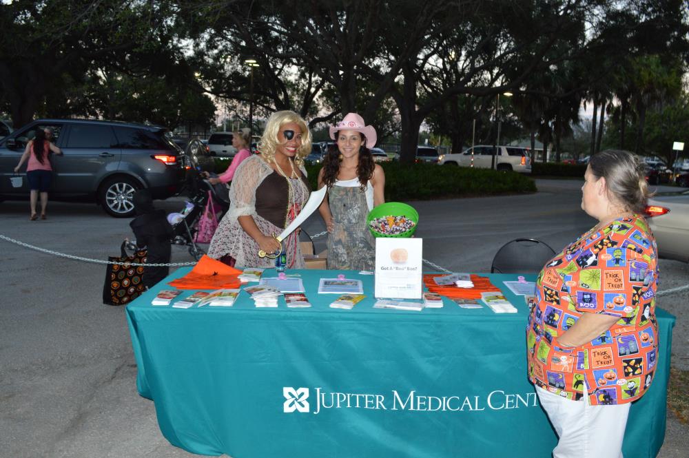 Woman dressed in pirate costume stands next to woman dressed in camo overalls and pink cowgirl hat behind Jupiter Medical Center information booth